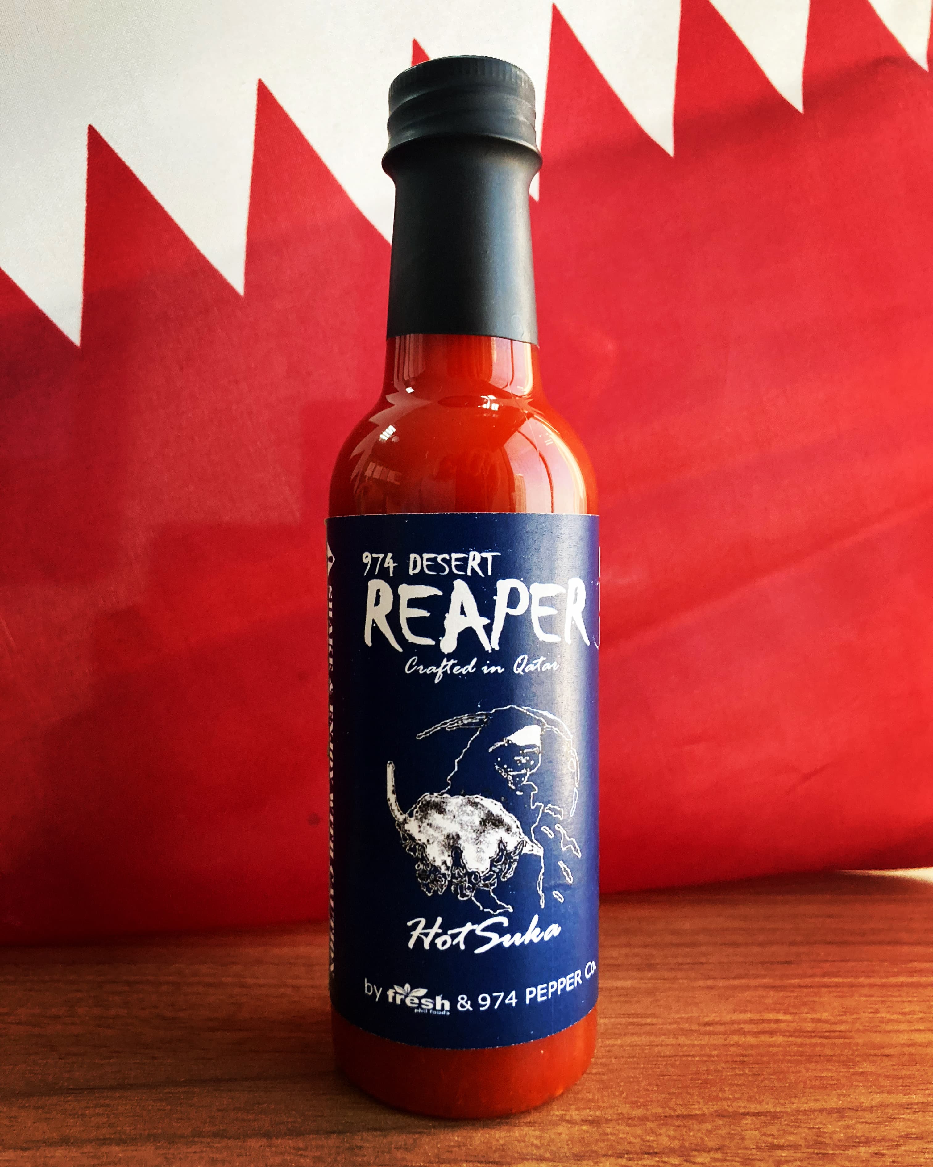 Bottle of red 974 Desert Reaper hot sauce on wood against the Qatari flag.